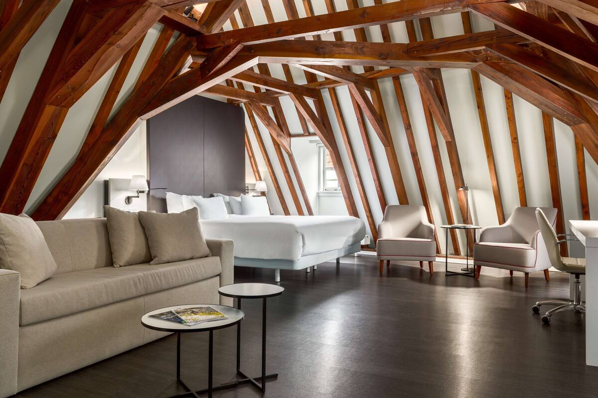 An attic bedroom with a high ceiling, exposed wooden beams, a bed, a sofa, a desk, and a chair.