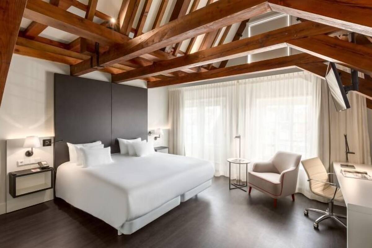 A modern hotel room with a large bed, a desk, a chair, and a window with curtains.
