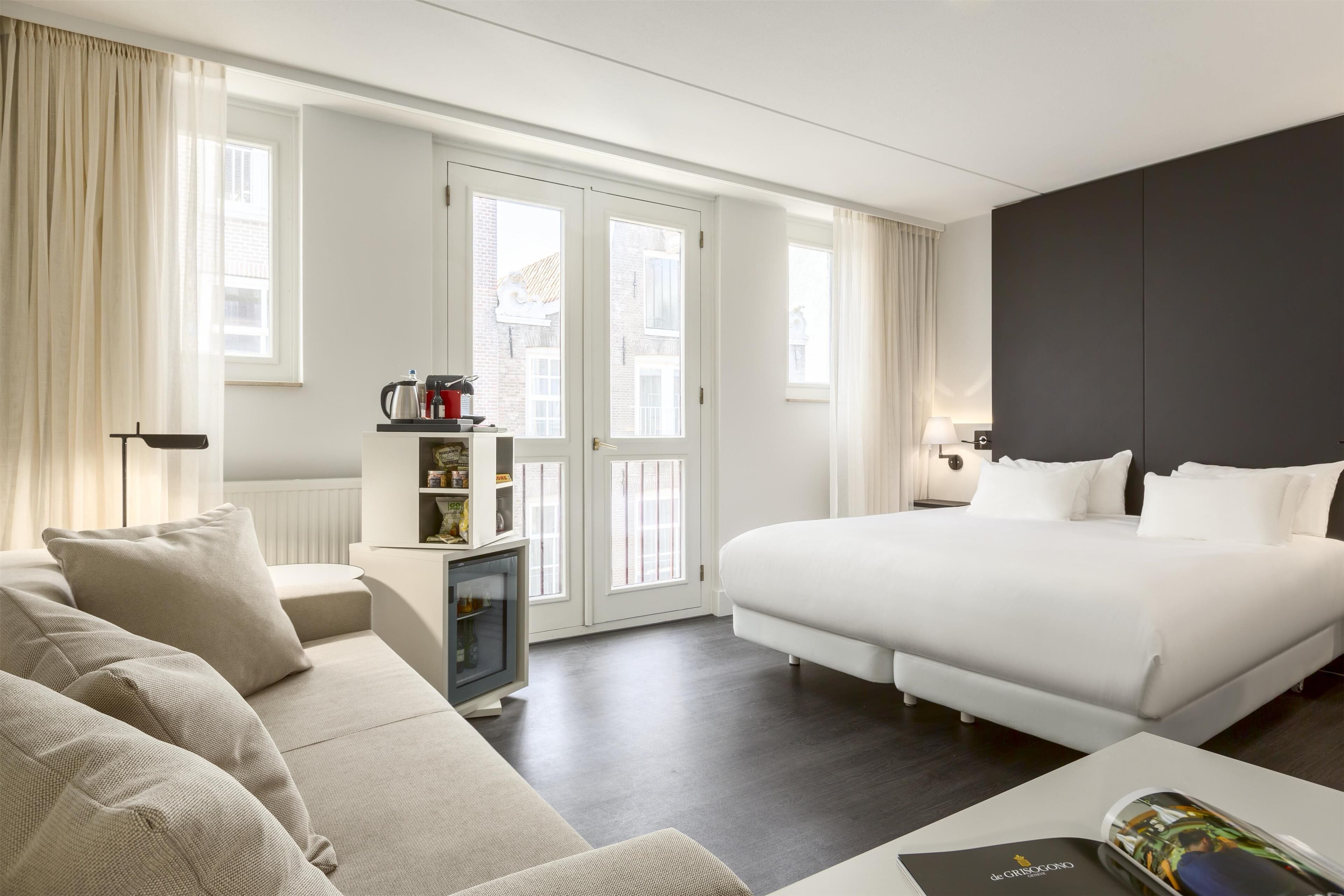 Ein modernes Hotelzimmer mit einem großen Bett, einer Couch, einem kleinen Beistelltisch und Blick auf die Stadt durch die Fenster.