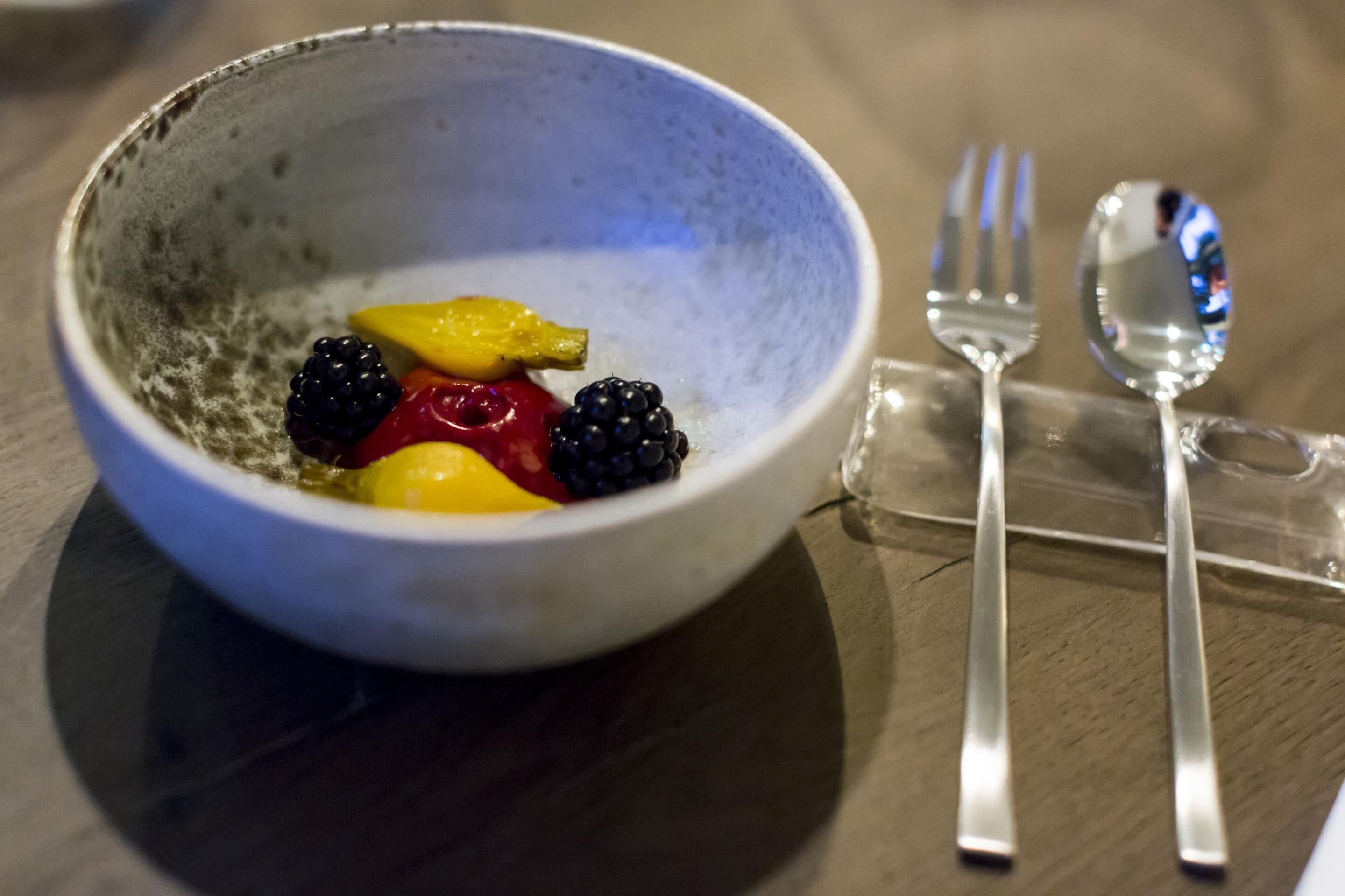 Un bol de fruta sobre una mesa, con un tenedor y una cuchara a un lado.