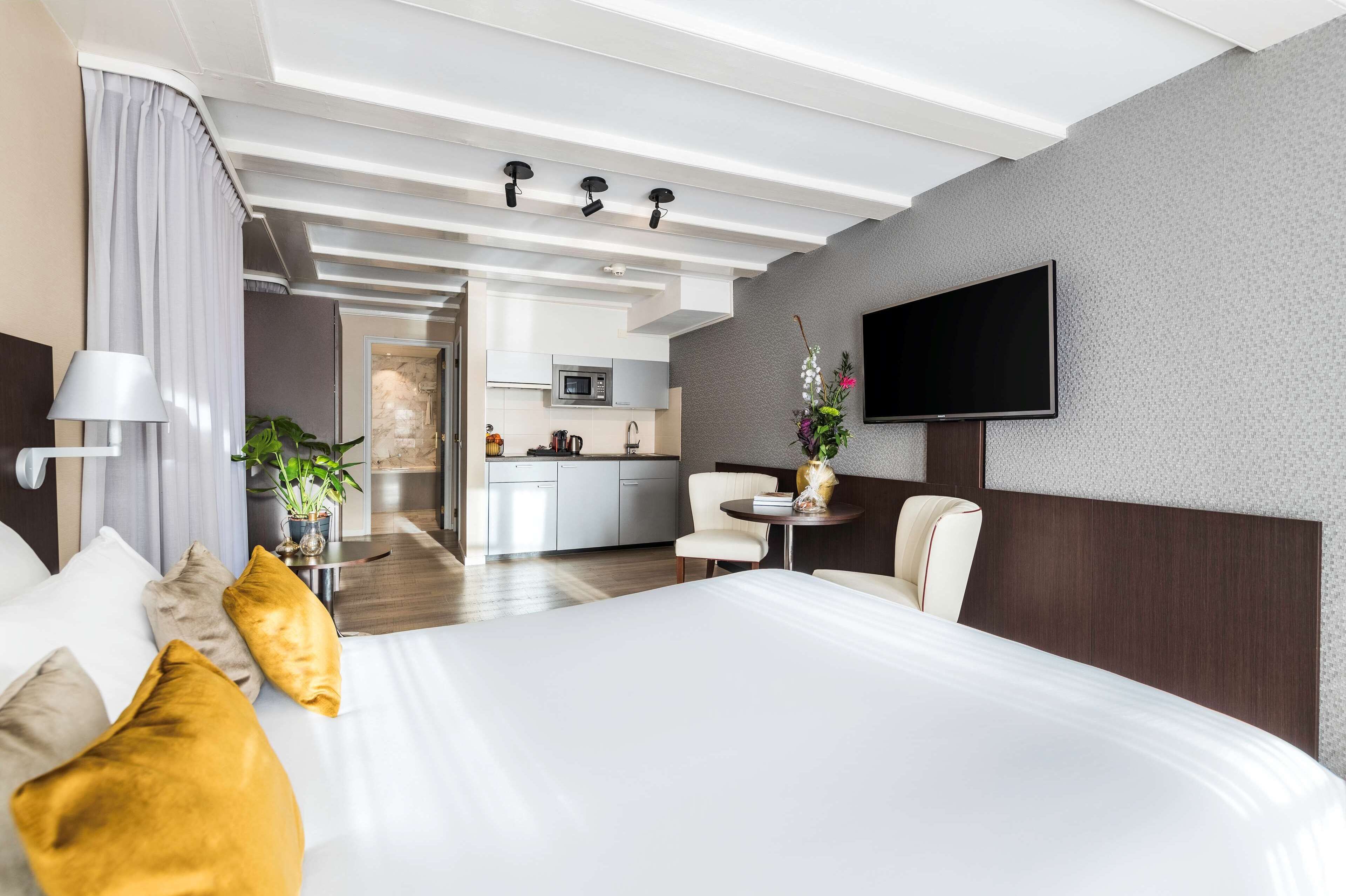 A hotel room with a large bed, a TV mounted on the wall, a small table with a vase, and a view of the kitchen and living area.