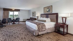 A hotel room with a bed, bedside table, lamp, chair, desk, and a view of the outdoors. - Shilo Inn Suites Idaho Falls (Idaho Falls)