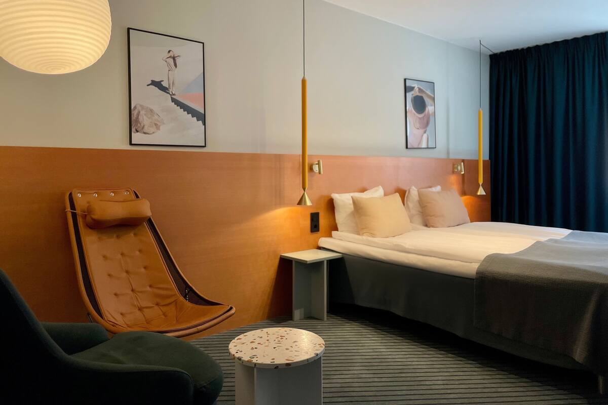 A modern hotel room with a large bed, a sitting area with a chair, a small table, and artwork on the walls.