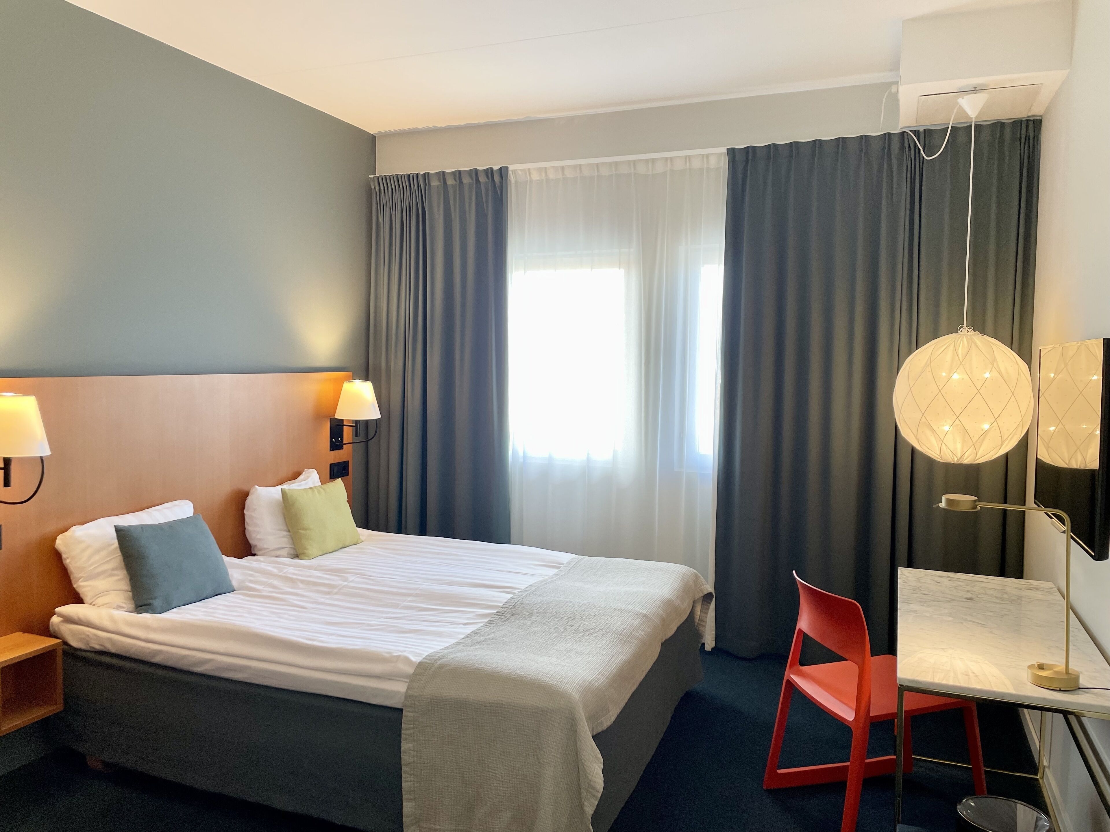Ein Hotelzimmer mit Bett, Schreibtisch, Stuhl, Lampe und einem großen Fenster mit Vorhängen.