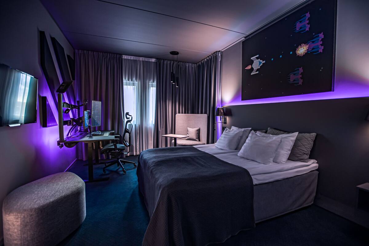 A modern hotel room with a large bed, a desk with a computer, a chair, and a TV.