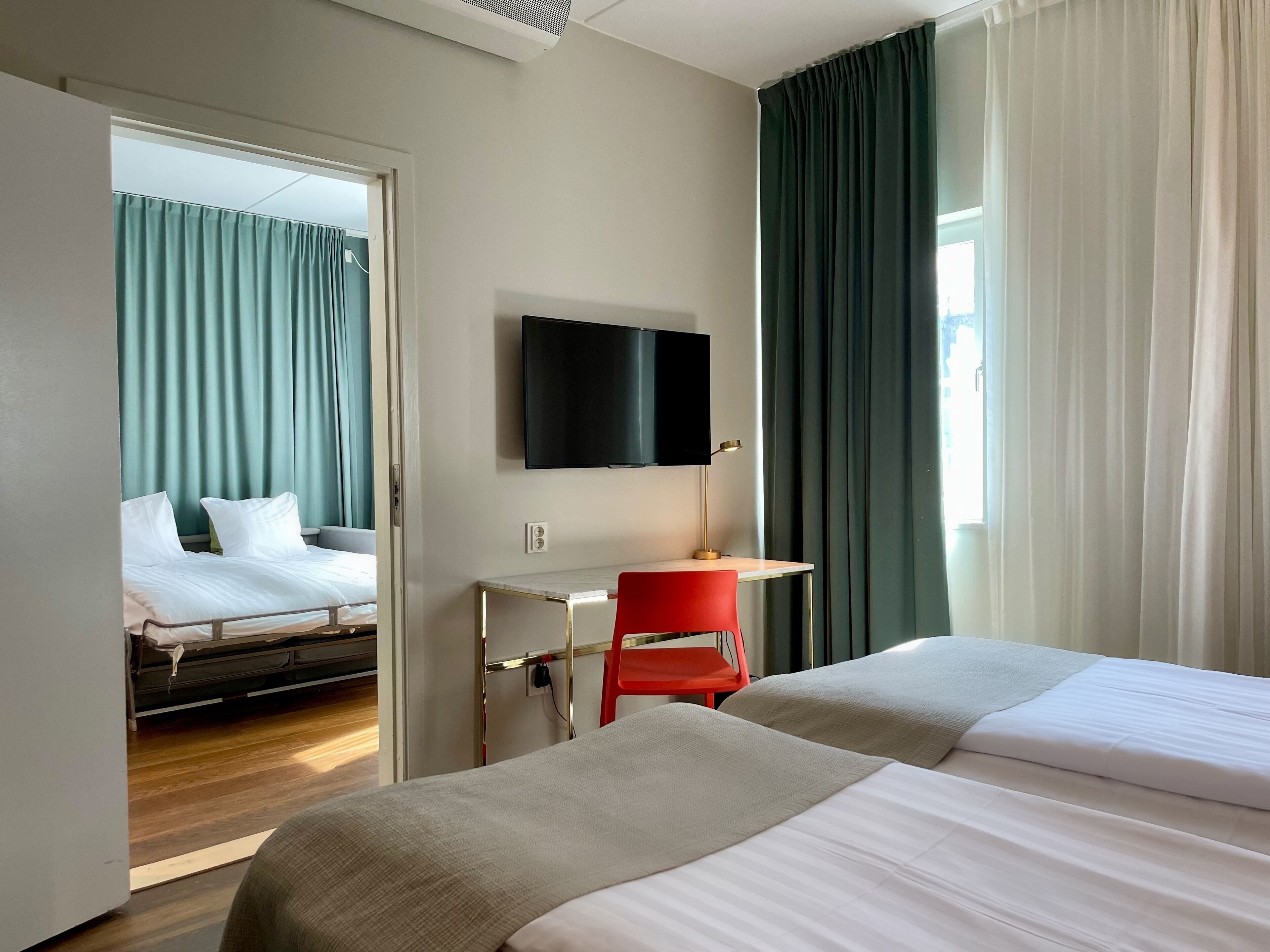 A hotel room with two beds, a desk, a TV, and a window with curtains.