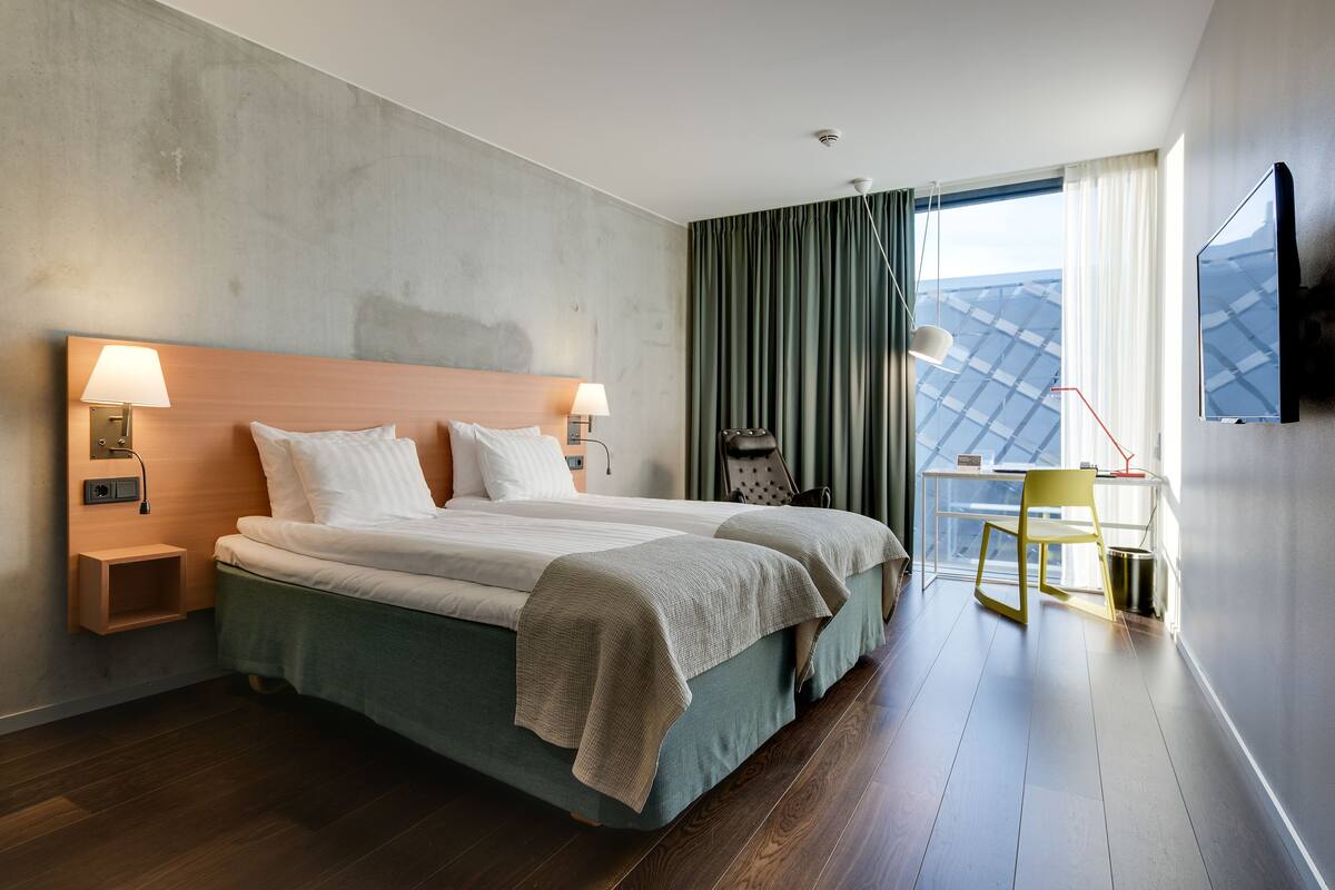 A modern hotel room with a large bed, two bedside tables with lamps, a desk with a chair, a TV mounted on the wall, and a view of the sky through the window.