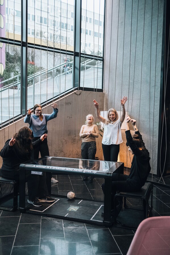A group of people in a modern indoor setting with large windows, a glass table, and a visible soccer ball.