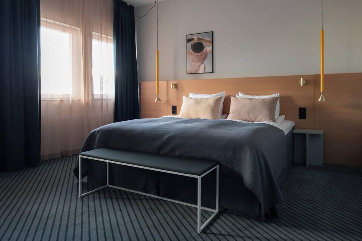 A hotel room with a large bed, a bedside table, a bench, and a window with curtains.