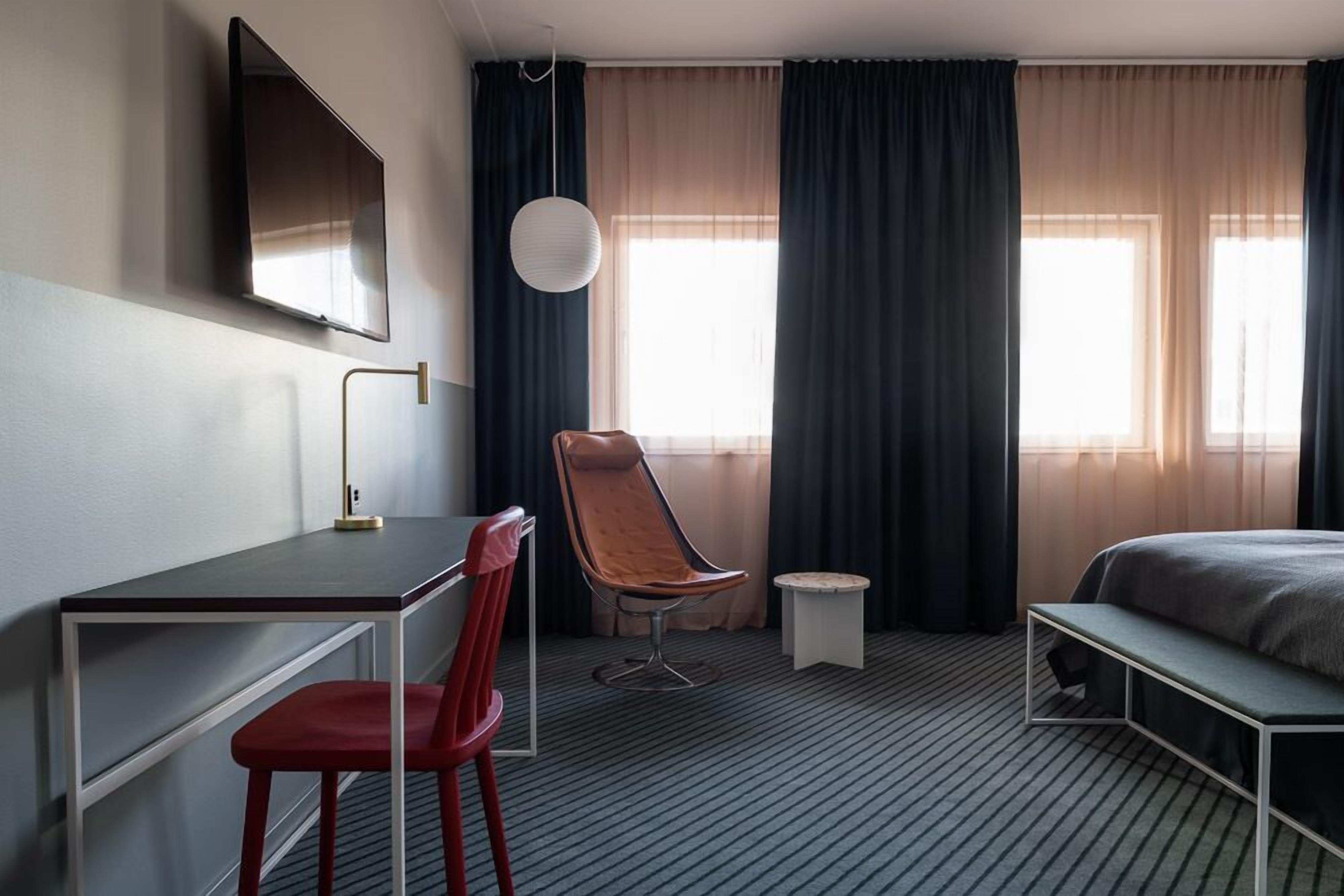 A hotel room with a bed, a desk with a chair, a TV mounted on the wall, and a window with curtains.