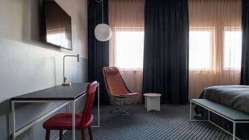 A hotel room with a bed, a desk with a chair, a TV mounted on the wall, and a window with curtains.