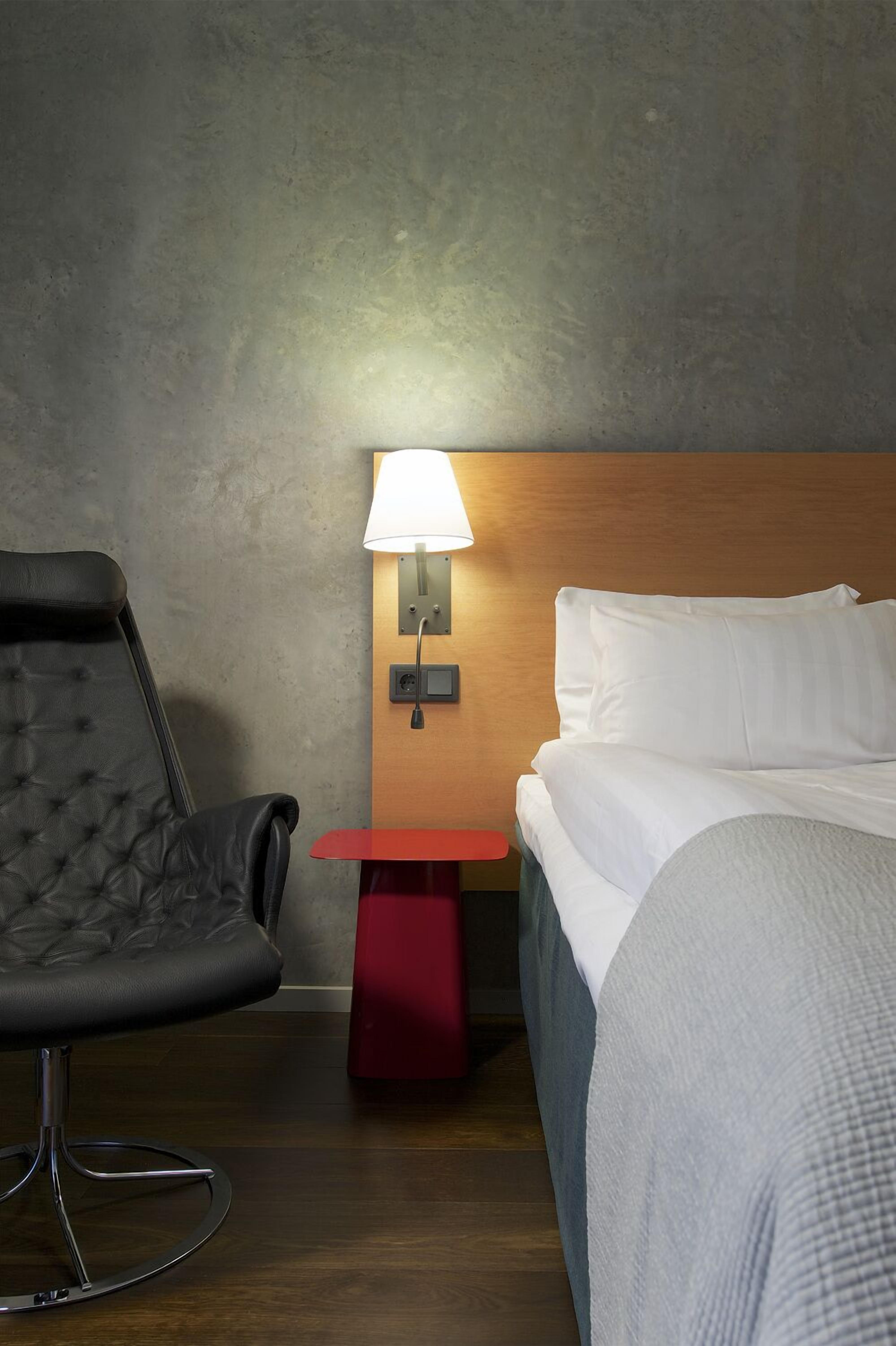 A hotel room with a bed, a chair, a nightstand with a lamp, and a wooden headboard.