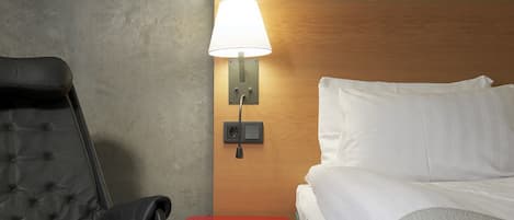 Habitación de hotel con cama, una silla, una mesita de noche con lámpara y cabecera de madera.