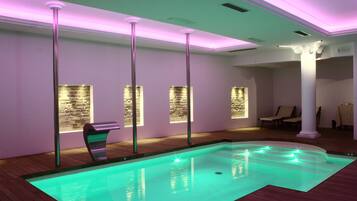 Una piscina interior moderna con suelo de madera y borde curvo, rodeada de columnas blancas y paneles de pared de piedra.