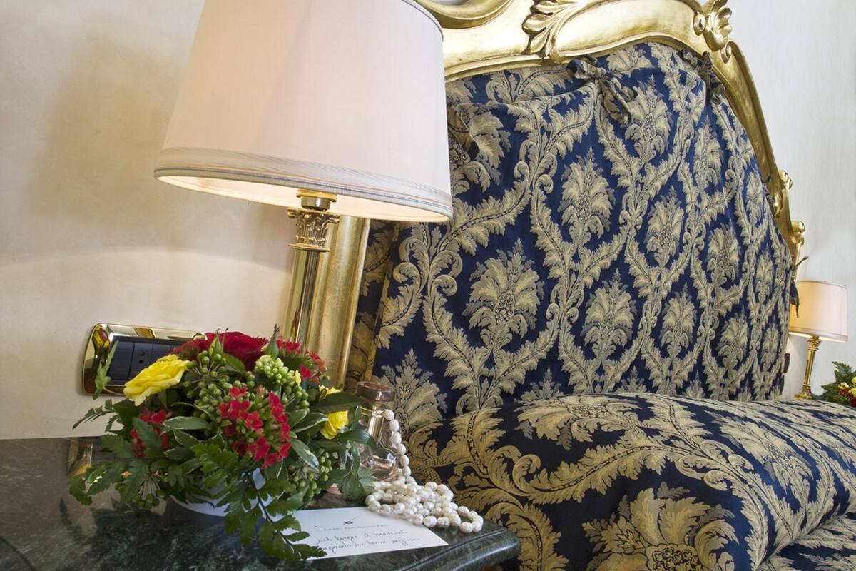 Una cama con cabecera dorada, una mesita de noche con lámpara y un arreglo floral.