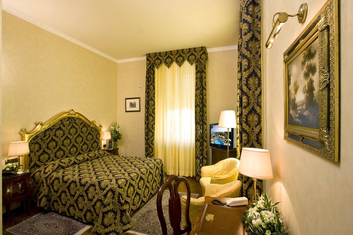 Habitación de hotel con una cama grande, una silla, un escritorio con lámpara, televisor y un cuadro en la pared.