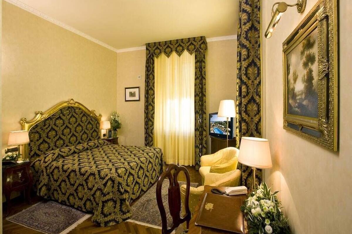 Una habitación de hotel con una cama grande, una silla, un escritorio con lámpara, un televisor y un cuadro en la pared.