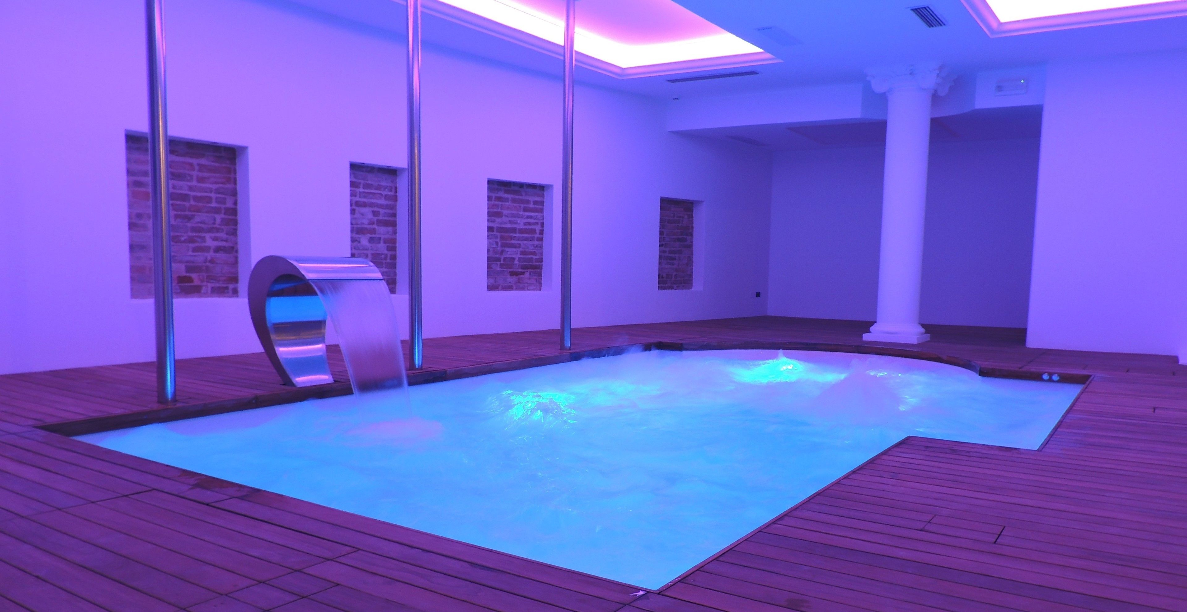 Una piscina interior moderna con cubierta de madera, paredes de vidrio y una iluminación azulada.