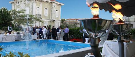 Un área de comedor junto a la piscina, con una fuente, mesas con comida y un edificio grande al fondo.
