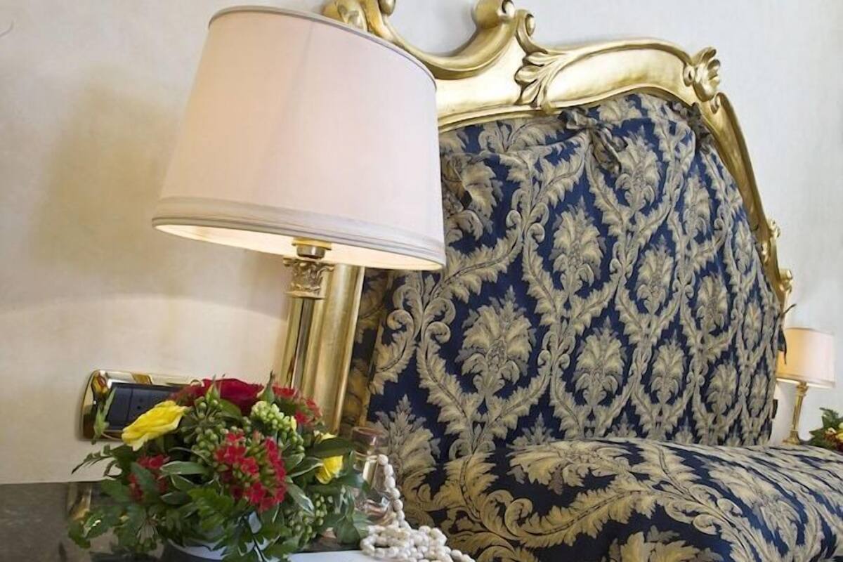 Una cama con cabecera azul y dorada, una mesita de noche con lámpara y un arreglo floral.