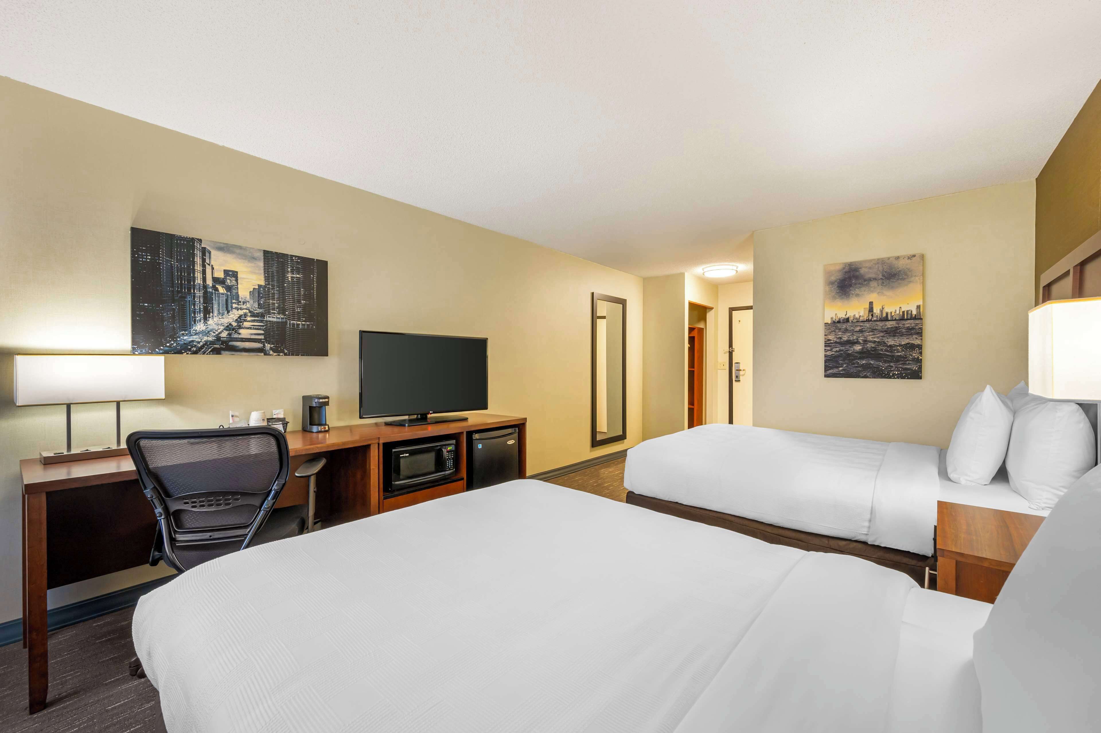 Ein Hotelzimmer mit einem großen Bett, einem Schreibtisch mit Fernseher, einem Sessel, einer Mikrowelle und einer Kaffeemaschine. An den Wänden hängen zwei Bilder.