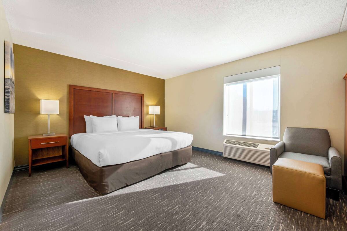 A hotel room with a large bed, two bedside lamps, a chair, a window, and a wall-mounted air conditioner.
