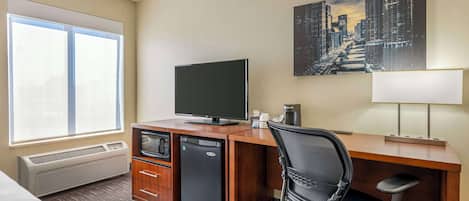 A hotel room with a bed, a desk with a television, a refrigerator, a chair, a window with blinds, and a cityscape painting on the wall.