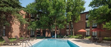 A rectangular swimming pool with a metal ladder, surrounded by lounge chairs and a brick building with windows.