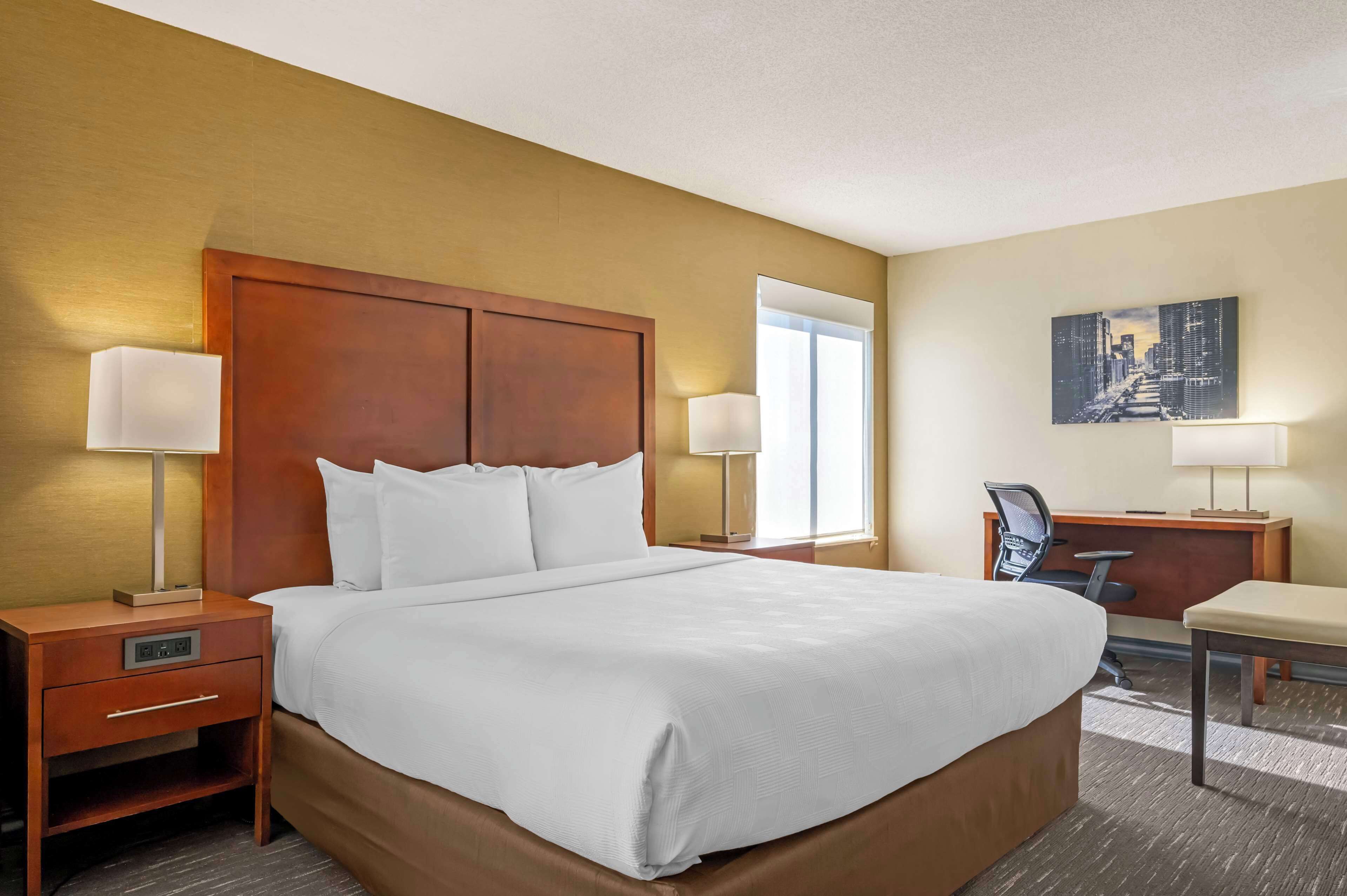 A hotel room with a large bed, two bedside tables with lamps, a desk with a chair, and a cityscape painting on the wall.