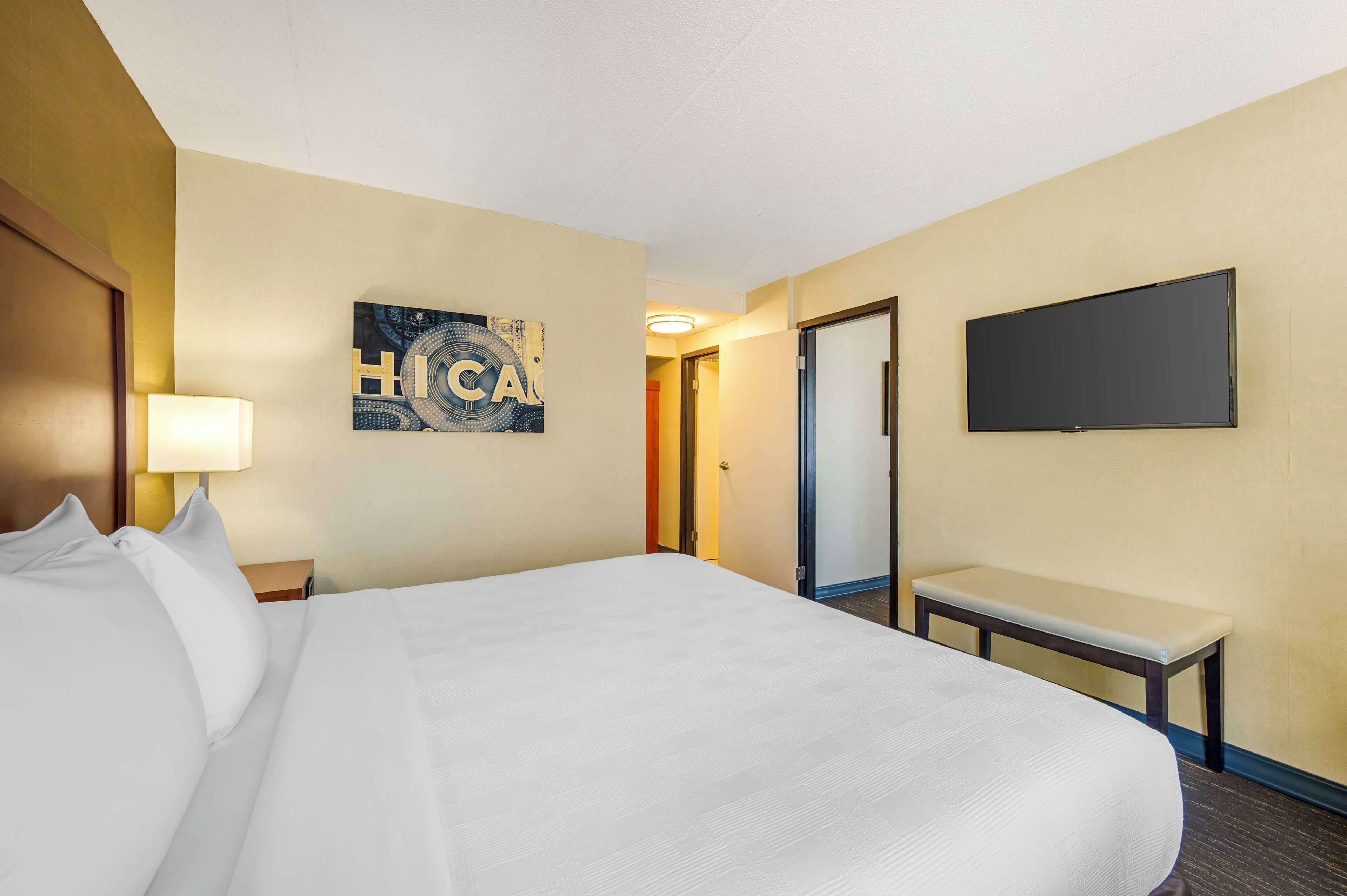 A hotel room with a large bed, a bedside table with a lamp, a flat-screen TV mounted on the wall, and a bench.