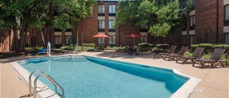 A rectangular pool with a metal ladder, surrounded by lounge chairs and umbrellas, with a brick building in the background.