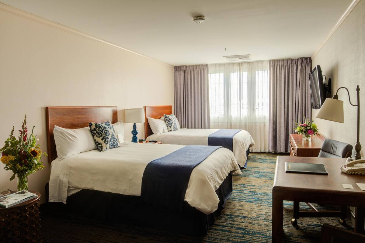 A hotel room with two beds, a desk, a chair, a TV, and a window with curtains.