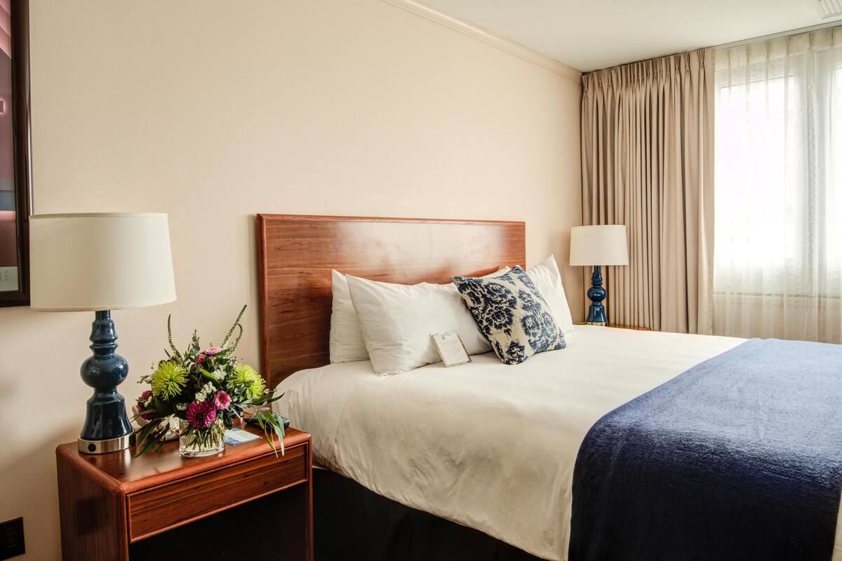 A hotel room with a wooden headboard, a large bed with white and blue patterned pillows, a dark blue blanket, a nightstand with a lamp and a vase of flowers.