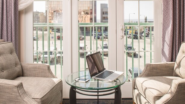 A room with two armchairs, a glass table with a laptop, and a view of a cityscape through a large window. - The Voyager Inn (Anchorage)