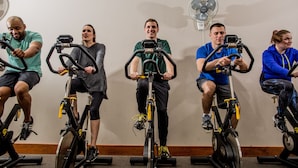 Um grupo de pessoas pedalando em bicicletas ergométricas numa sala com parede branca e um ventilador de pé.