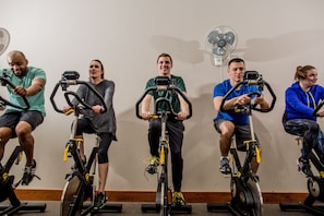 A group of people on stationary bikes in a room with a white wall and a standing fan. - The Voyager Inn (Anchorage)