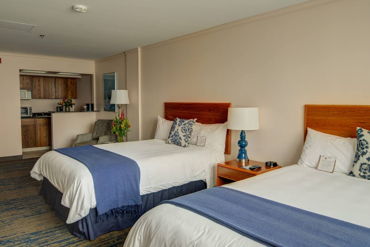 A hotel room with two beds, a wooden headboard, a desk with a lamp, and a kitchenette in the background.