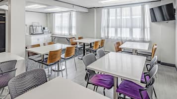 Un comedor moderno con mesas y sillas blancas, una zona de cocina con electrodomésticos y ventanas amplias con cortinas ligeras.