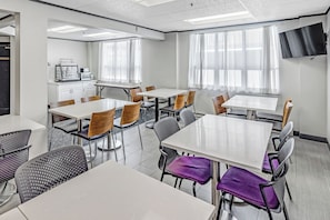 A modern dining area with white tables and chairs, a kitchen area with appliances, and large windows with sheer curtains.