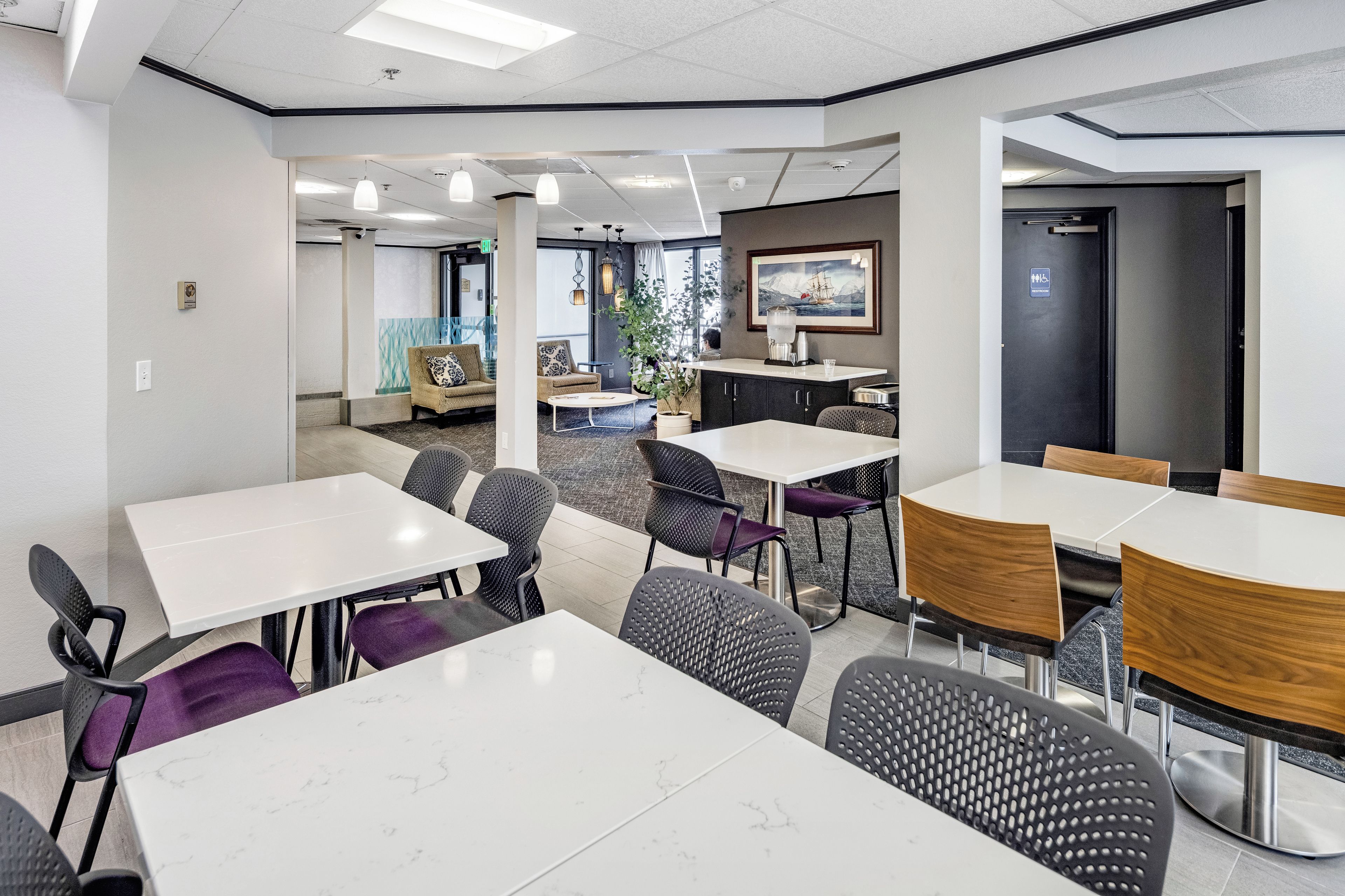 A modern indoor space with multiple seating areas, including tables and chairs, and a large window providing natural light.