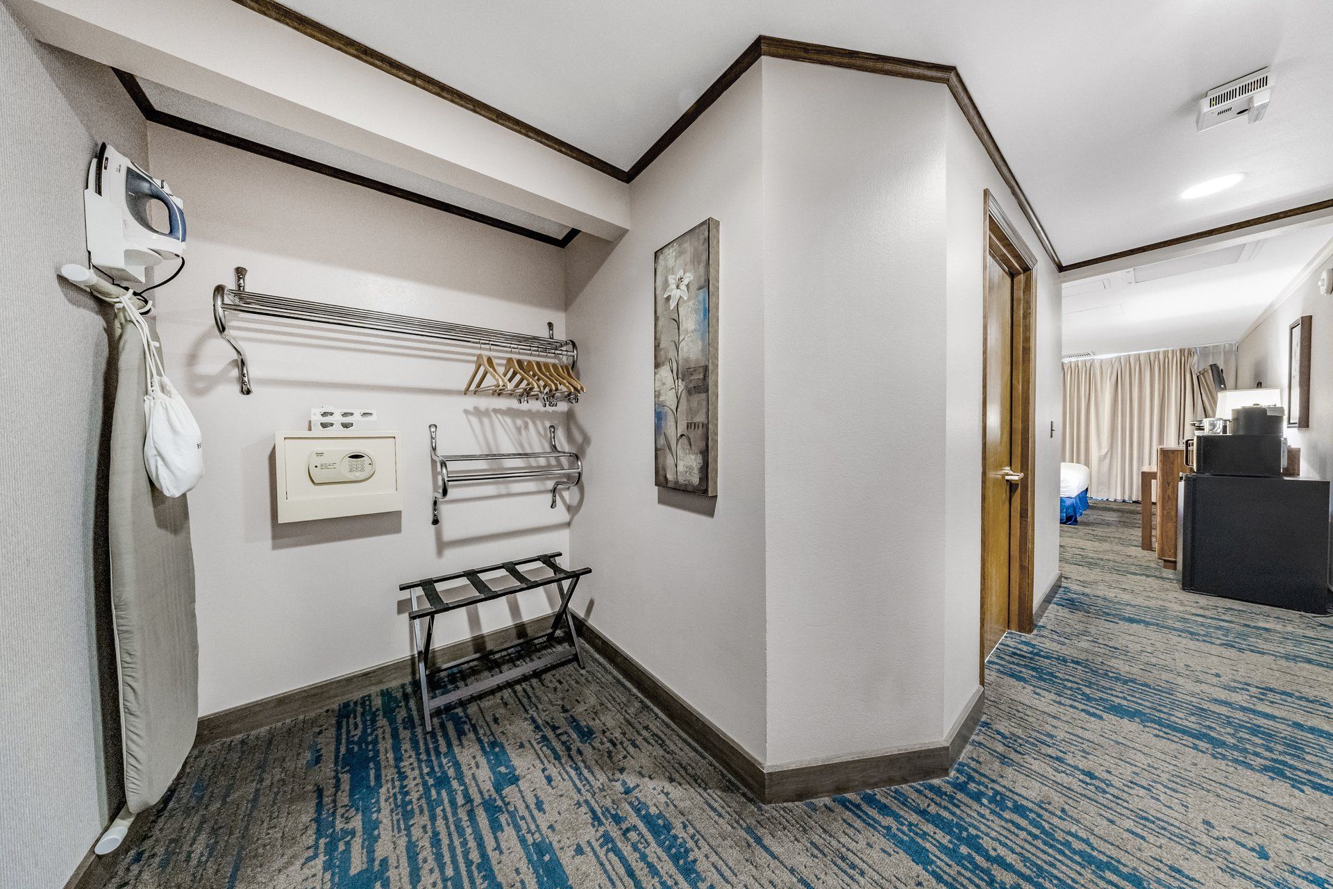 Pasillo de hotel con perchero, toallero, un cuadro y una puerta que da a un dormitorio.