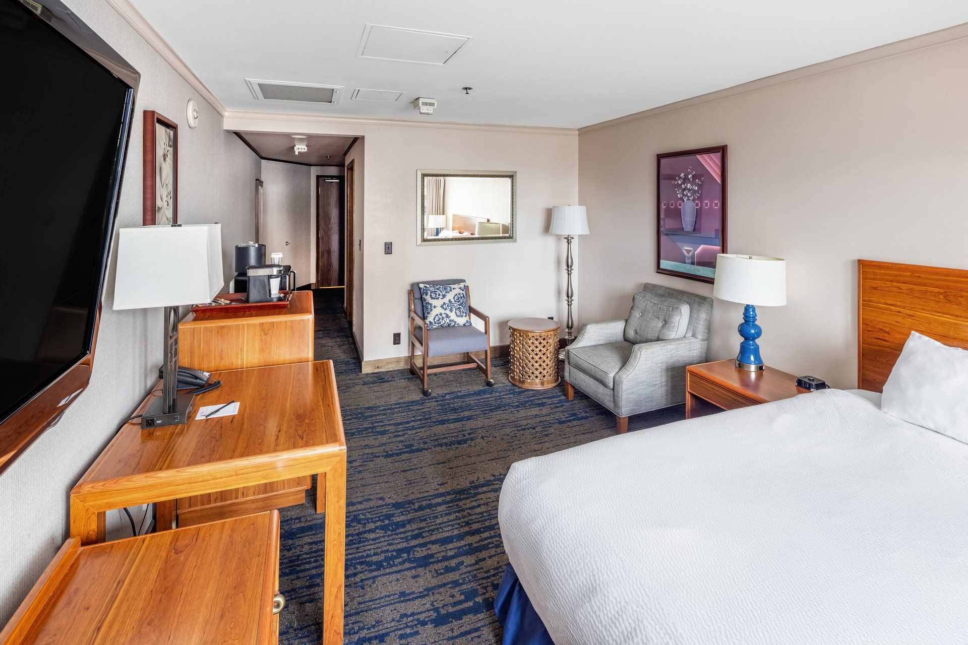 A hotel room with a bed, a desk, a chair, a TV, a lamp, and a painting on the wall.