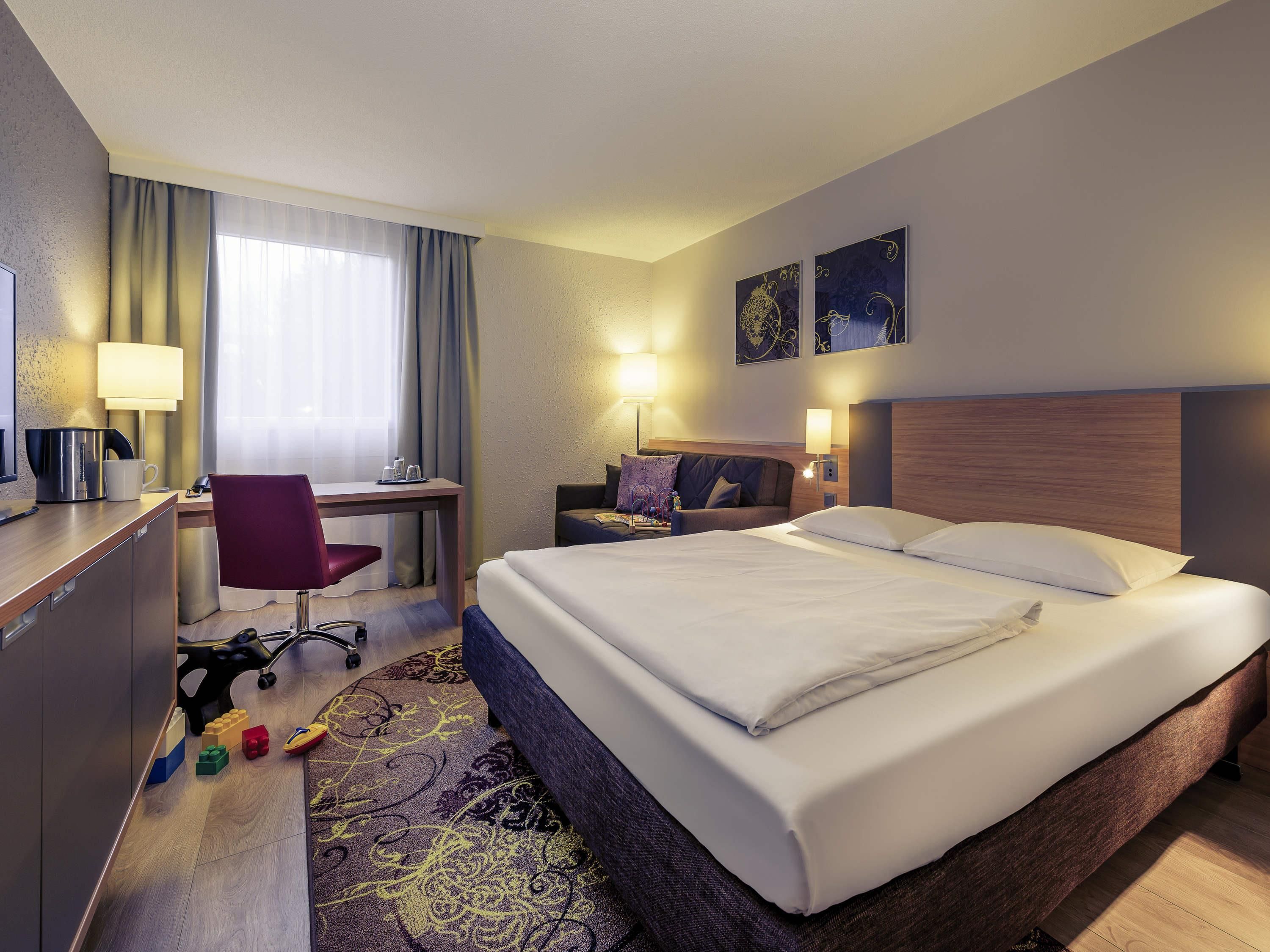 A hotel room with a bed, desk, chair, and a small table with a lamp.