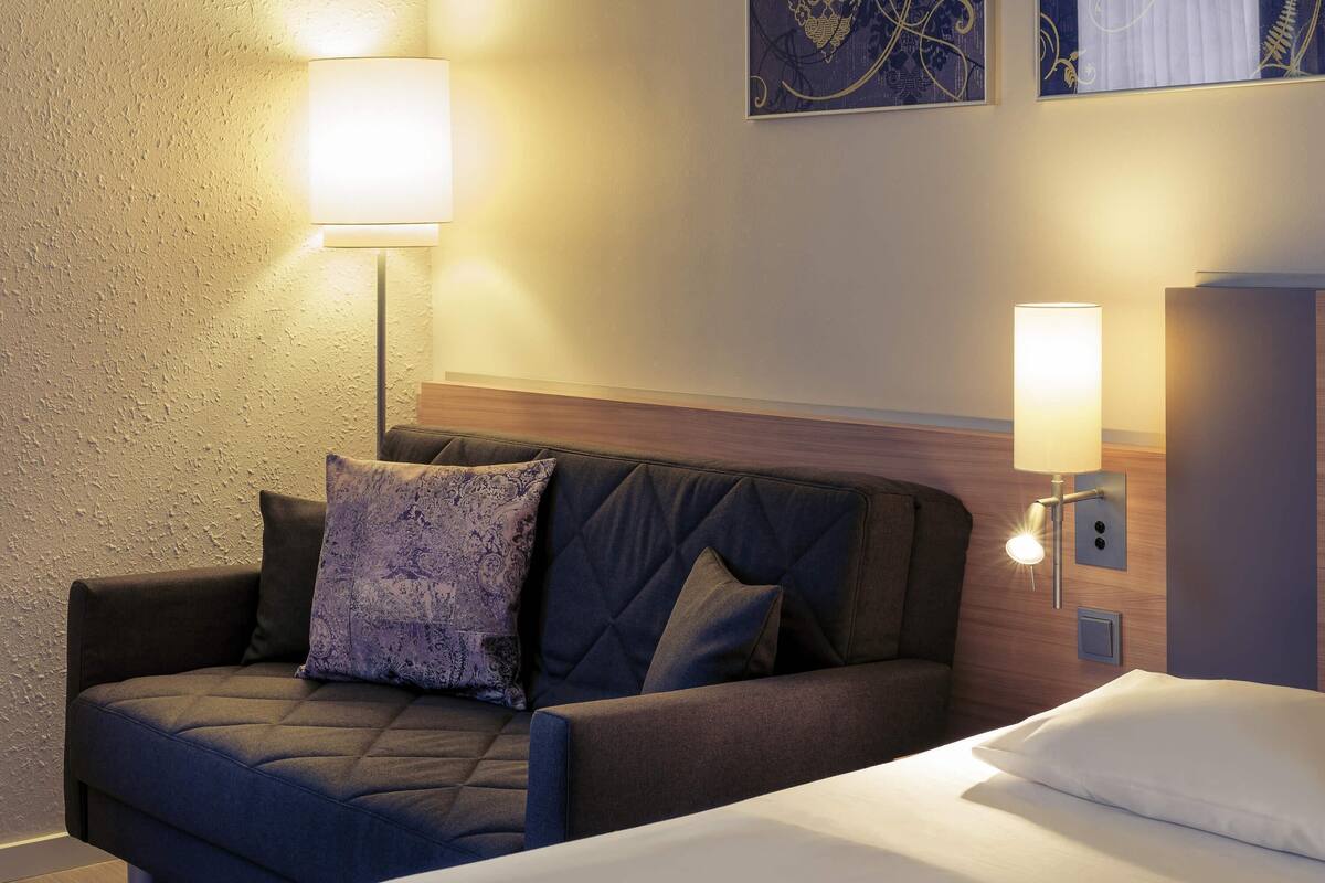 Una habitación de hotel con cama, sofá, lámpara de pared y un cuadro.