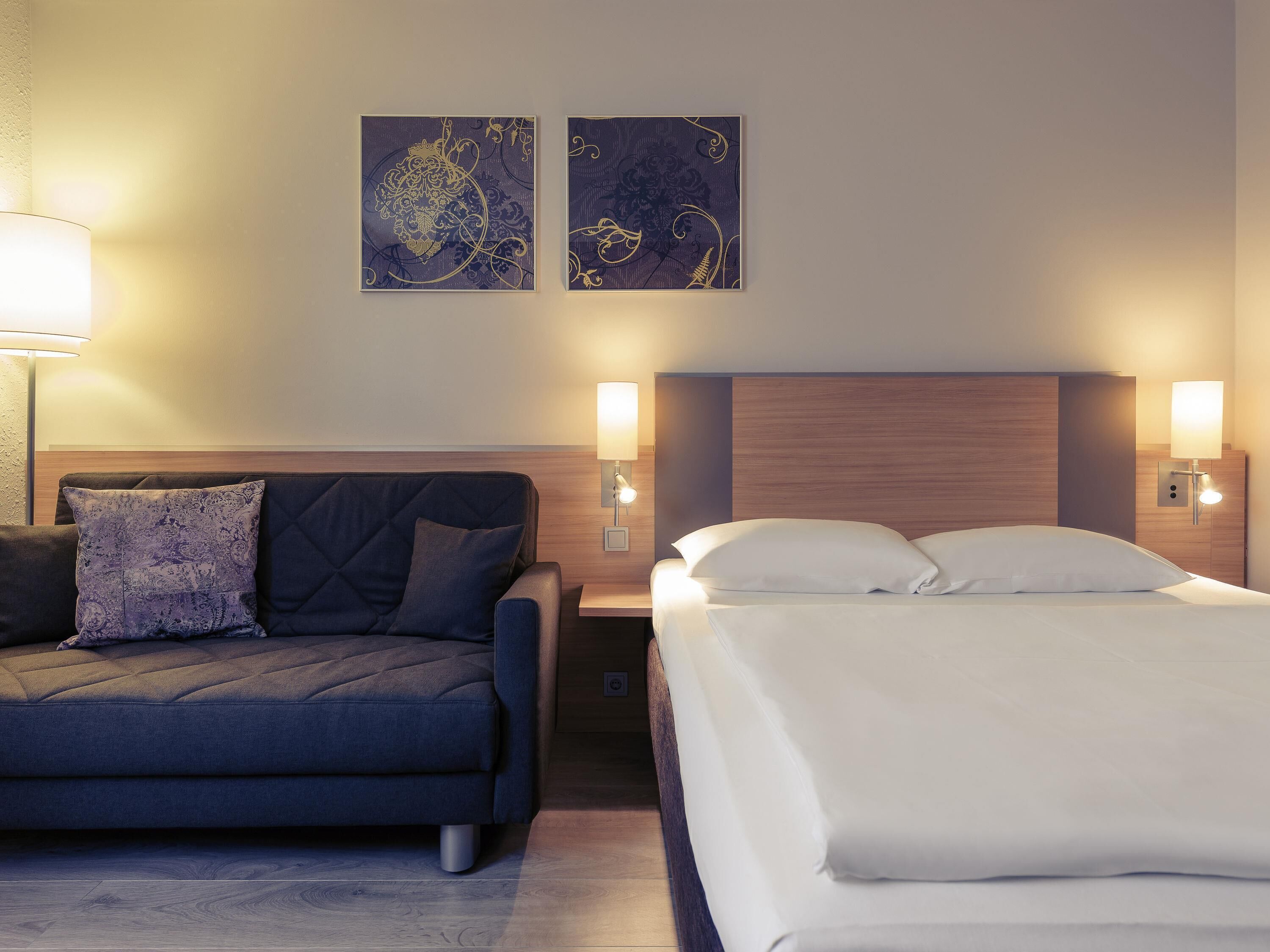 A hotel room with a bed, a sofa, two wall-mounted lamps, and two framed artworks.