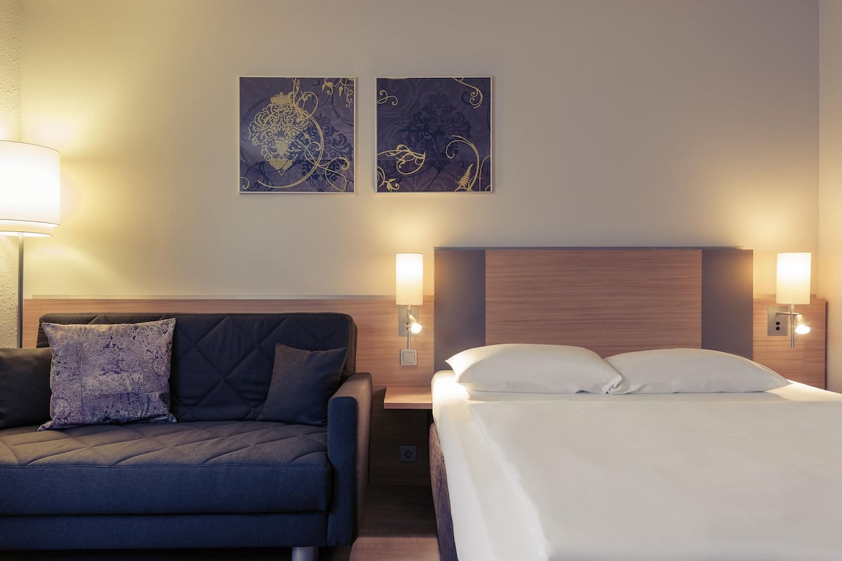 Une chambre dâhĂŽtel comprenant un lit, un canapĂ©, deux lampes fixĂ©es au mur et deux Ćuvres dâart encadrĂ©es.