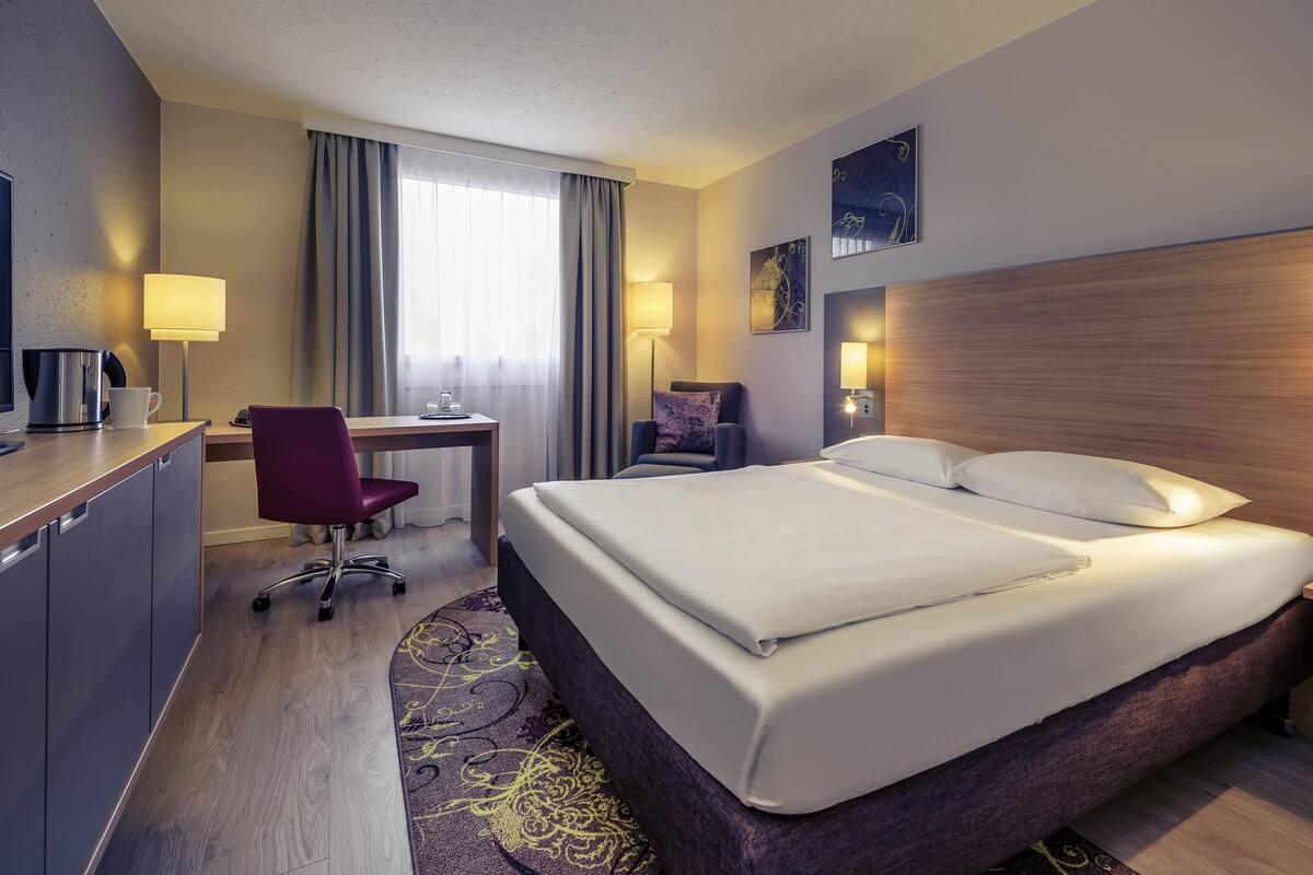 A hotel room with a bed, desk, chair, and a small table with a lamp.