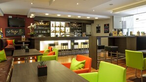 A modern bar area with a counter, seating with bright green chairs, and a variety of bottles and glasses on shelves.