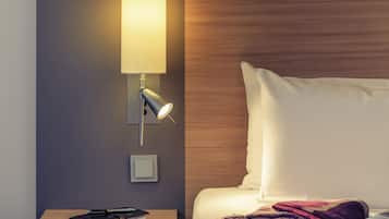 A neatly made bed with a white pillow and a purple blanket, a bedside table with a book and a pen, and a wall-mounted lamp with a light on.