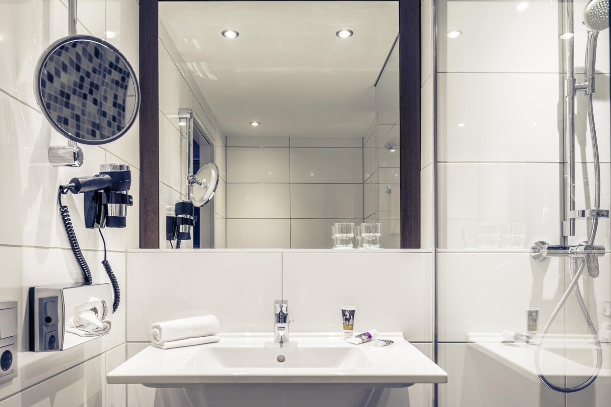 A modern bathroom with a large mirror, a hairdryer, a towel, a sink, and a shower.